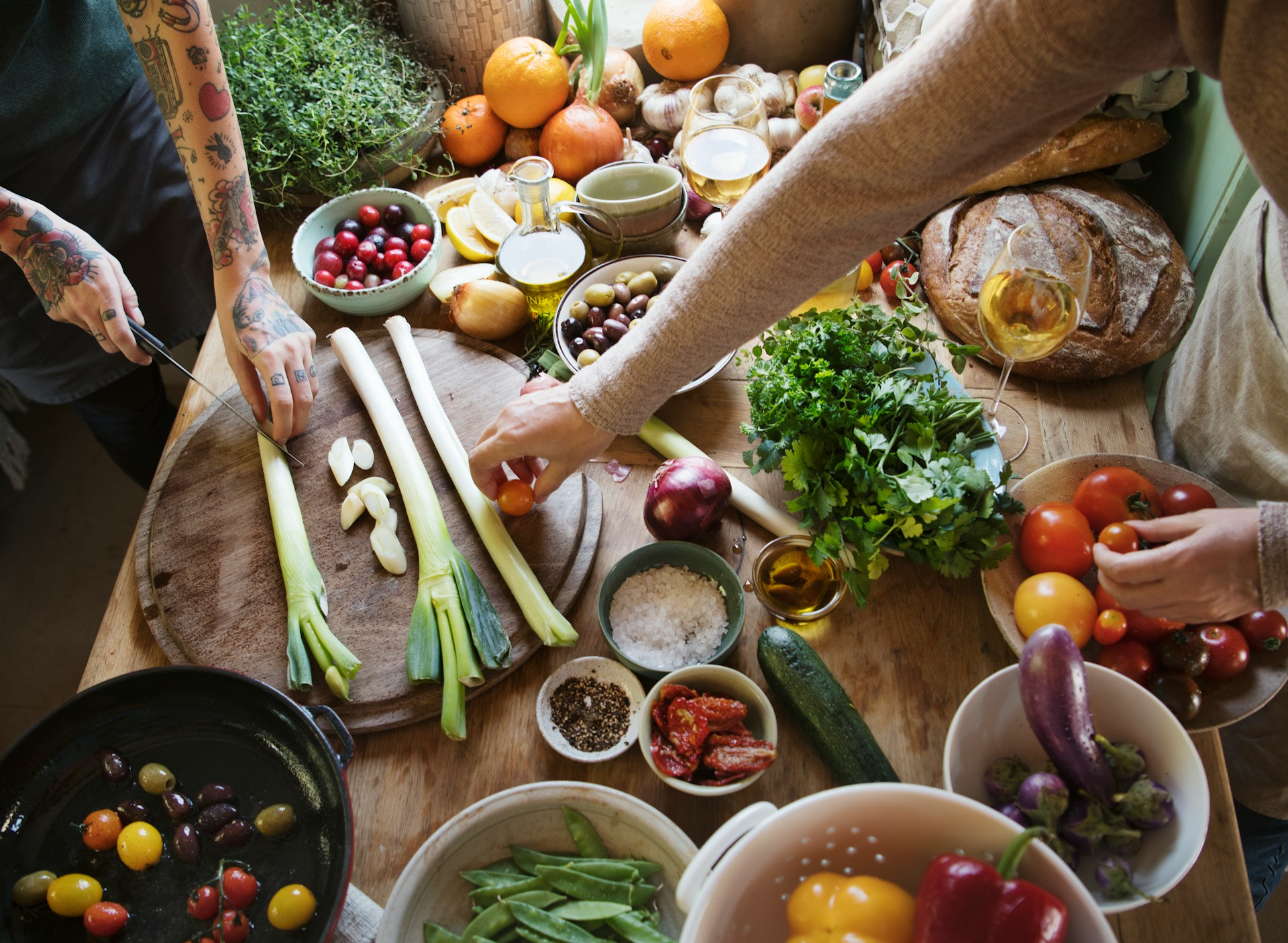 A wooden counter with a variety of fresh vegetables laid across it, with some small dishes of spices and a loaf of bread. Two sets of hands are seen: one set on the left is chopping leek, and the set on the right is picking up ingredients.